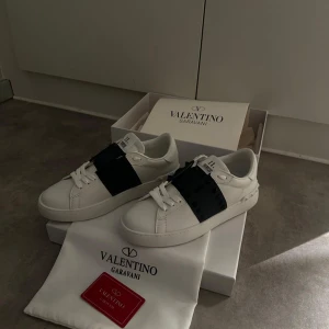 Valentino Garavani sneakers i vitt och mörkblått  - Säljer ett par vita Valentino Garavani sneakers med en bred mörkblå rem över vristen dekorerad med mörkblåa nitar. Skorna har klassisk snörning och en platt sula. Kommer med originalpåse och låda. Skorna är sprillans nya och är i topp skick. Priset kan diskuteras vid snabbaffär🤩.Hör av vid minsta fundering 🤝.