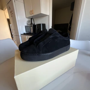  svarta sneakers från Axel Arigato - Säljer ett par helsvarta sneakers från Axel Arigato med minimalistisk design. Skorna har en låg profil, rund tå och är tillverkade i mocka med diskret logga på sidan. Perfekta för dig som gillar stilrena och cleana skor.