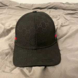 Svart keps med logotypmönster - Snygg svart keps med diskret logotypmönster och klassiska ränder i grönt och rött på sidan. Perfekt för en stilren look.