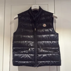 Navy Moncler gui väst - Säljer en marinblå dunväst från Moncler med hög krage och två dragkedjefickor framtill. Västen är storlek 3 vilket motsvarar M-L. Passar perfekt till mig som vanligtvis bär M. Den är använd vid ett få tillfällen och stått mest i garderob därav säljes den. Västen har Moncler-logga på bröstet och stängs med dragkedja. Perfekt för lager-på-lager och stilren look. Hör av er vid frågor!