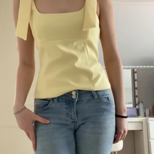 Gul linnetopp med rosettband - Säljer en söt gul linnetopp med breda axelband som knyts till rosetter på axlarna. Toppen har fyrkantig ringning och figurnära passform. Perfekt för varma dagar! Helt ny! Lapp är kvar 