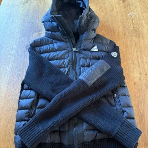 Svart cardigan från Moncler - Säljer en snygg svart pufferjacka från Moncler med stickade ärmar och huva. Jackan har dragkedja och ett Moncler-märke på armen. Ställ frågor vid minsta funderingar