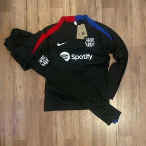FC Barcelona svart långärmad träningströja från Nike - Säljer en svart långärmad FC Barcelona träningströja från Nike med klubbmärke, vit Nike-logga och Spotify som sponsor. Tröjan har dragkedja vid halsen och röda samt blå detaljer på axlarna. Materialet är funktionsmaterial.