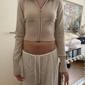 Beige croppad hoodie - Säljer en beige croppad hoodie med dragkedja framtill. Tröjan har långa ärmar och ribbade detaljer vid midjan och ärmsluten. Perfekt för en avslappnad stil.