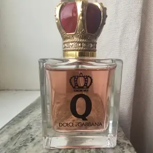 Elegant parfymflaska från Dolce & Gabbana med en kunglig krona som lock. Flaskan är genomskinlig med en rosa vätska inuti och har en gyllene krona med röda detaljer. Perfekt för den som vill ha en lyxig doftupplevelse. Ej använd 50ml köptes för 1300kr