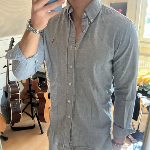 Grå skjorta från Ralph Lauren - Snygg grå skjorta från Ralph Lauren med broderad logga på bröstet. Skjortan har en klassisk krage och knappar framtill. Perfekt för en stilren look.