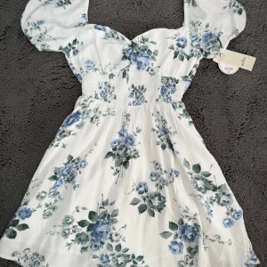 Hollister klänning - Vit klänning med blått blommönster från Hollister. Den har puffiga korta ärmar och en smockad midja för en snygg passform. Den har inbyggda shorts och fickor. Klänningen är öppen i ryggen och har en jätte söt knytning. Även en fin urringningen i fram med vridet tyg🤩✨💗