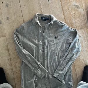Grå skjorta från Ralph Lauren - Säljer en stilren grå skjorta från Ralph Lauren i storlek XS. Skjortan är långärmad och har knappar framtill. Den är tillverkad i ett lätt och bekvämt material med en broderad logotyp på bröstet. Den behöver strykas som ni ser på bilden annars är din i bra skick! Skriv gärna om ni har några funderingar 🤗🤗🤗