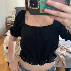 Svart blus från Uniqlo - Säljer en söt svart blus från Uniqlo med korta puffärmar och elastisk midja. Blusen har ett diskret hålmönster och en bekväm passform. Perfekt för en stilren look!