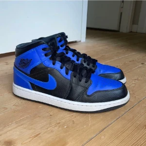 Nike Air Jordan 1 Mid i svart och blå - Säljer ett par Nike Air Jordan 1 Mid i färgerna svart och hyper royal blå. Skorna har en klassisk design med snörning och en vit sula. Perfekta för sneaker-entusiaster som vill ha en stilren look.  Jordan 1 mid hyper royal Storlek 10.5US=44.5EU Köpt pa Soleplus