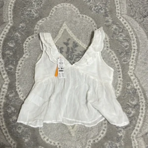 Vit volangtopp kosta 200kr och säljer för 130 man kan dra ner priset  - Säljer en söt vit volangtopp med v-ringning och volangdetaljer. Toppen är ärmlös och har en luftig passform, perfekt för varma dagar. Den är gjord i ett lätt och bekvämt material 