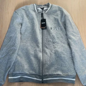 Grå cardigan från Boss - Snygg grå cardigan  från Boss med dragkedja framtill och ribbade muddar. Jackan har en stilren design med broderad logga på bröstet och kontrasterande ränder vid ärmslut och nederkant.Helt ny 🆕 
