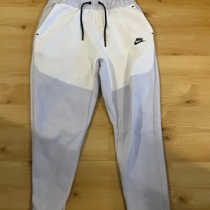 Vita mjukisbyxor från Nike - Säljer ett par vita mjukisbyxor från Nike med svart logga på framsidan. Byxorna har en elastisk midja med snörning och sidofickor. Perfekta för en avslappnad stil. OBS FLÄCKAR PÅ NEDRE DELEN MEN INGET SOM SYNS NÄR MAN HAR DOM PÅ SIG