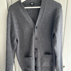 Grå kofta från Uniqlo - Säljer en stilren grå kofta från Uniqlo med knappar framtill och två praktiska fickor. Koftan har en v-ringad design och är perfekt för kyligare dagar. Passar både till vardags och mer uppklädda tillfällen.