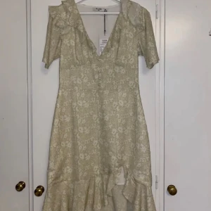 Blommig klänning från NA-KD - Säljer en vacker blommig klänning från NA-KD i ljusgrön och vit. Klänningen har en v-ringad design med volangdetaljer och korta ärmar. Perfekt för sommarens alla tillfällen! Prislapp kvar