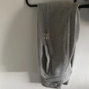 Grå mjukisbyxor från Adidas - Säljer ett par grå mjukisbyxor från Adidas med elastisk midja och sidofickor. Perfekta för en avslappnad stil eller träning. Adidas-loggan är tryckt på ena benet. 
