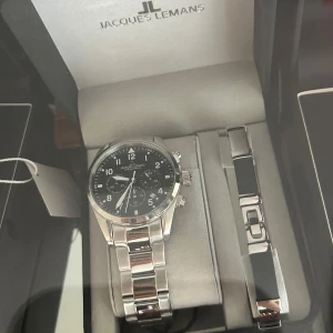 Jacques Lemans klocka i silver (oanvänd) - Elegant klocka från Jacques Lemans med en stilren silverfärgad länkarmband och svart urtavla. Klockan har tydliga siffror och visare, samt tre mindre urtavlor för extra funktioner. Perfekt för den som söker en klassisk och tidlös accessoar. Vattentät 100m