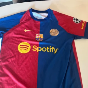 FC Barcelona fotbollströja - Säljer en FC Barcelona fotbollströja från Nike med korta ärmar. Tröjan har ett delat mönster i rött och blått med gula detaljer. Den har klubbens emblem och sponsorloggor på framsidan samt spelarnamn och nummer på baksidan.