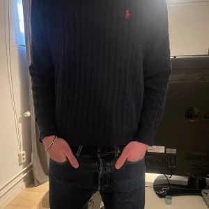Mörkblå stickad tröja från Ralph Lauren (L) - Säljer en stilren mörkblå stickad tröja från Ralph Lauren med det klassiska broderade logotypen på bröstet. Tröjan har en ribbad struktur och långa ärmar, perfekt för kyligare dagar. Passar både till jeans och chinos för en avslappnad look.