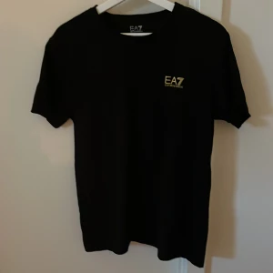 Svart t-shirt från EA7 Emporio Armani - Svart t-shirt från EA7 Emporio Armani med gul logga på bröstet. Klassisk passform och rund halsringning. Perfekt för en stilren och enkel look.