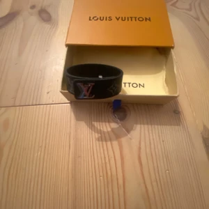 Svart armband från Louis Vuitton - Säljer ett svart armband från Louis Vuitton med klassiskt LV-logo i metall framtill. Armbandet har monogrammönster och är tillverkat i läder. Kommer med originalask, dustbag och äkthetskort. Tillverkat i Spanien. Allt i bilderna kommer med! Ge ditt pris!
