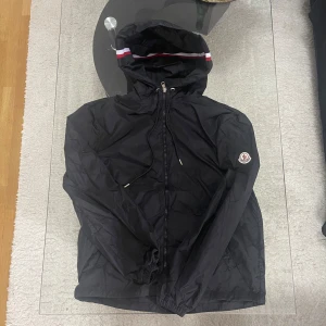 Svart vindjacka från Moncler med huva - Säljer en svart vindjacka från Moncler med huva och dragsko. Jackan har dragkedja framtill och Moncler-logga på ärmen. Huvan har en snygg detalj med röd och vit rand. Perfekt för blåsiga dagar. Lågare pris för dragkedjan är inte topp kvalitet längre där den kan fastna. Size 3 passar nu s-m för den är gammal