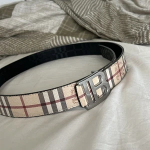 Burbery bälte  - Snyggt bälte med klassiskt rutigt mönster i beige, rött och svart. Bältet har ett stilrent metallspänne med bokstäver. Perfekt för att ge en touch av elegans till din outfit.