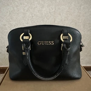 Svart handväska från Guess - Snygg svart handväska från Guess med guldfärgade detaljer och dragkedja. Väskan har två handtag och är perfekt för att bära dina nödvändigheter med stil. Guess-loggan pryder framsidan och ger en lyxig touch.