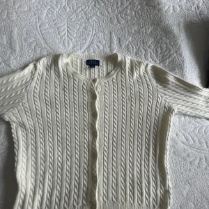 Vit kabelstickad kofta från Polo Ralph Lauren - Säljer en klassisk vit kabelstickad kofta från Polo Ralph Lauren med rund halsringning och knappar framtill. Tröjan har långa ärmar och en liten broderad logga i ljusrosa på bröstet. Perfekt att slänga över en topp eller klänning. Koftan är i superfint skick och är knappt använd! Koftan har inget tecken på användning eftersom det inte är några defekter/fläckar eller nopper!🥰 nypris ca: 1600kr