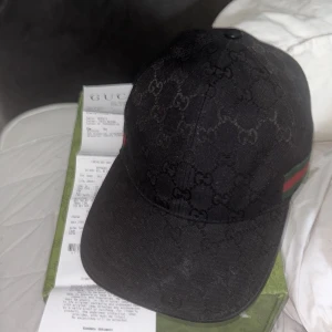 Svart keps från Gucci med GG-mönster - Säljer en svart keps från Gucci med klassiskt GG-mönster och grönt/rött band på sidan. Kepsen har böjd skärm och justerbar passform. Perfekt för dig som vill ha en stilren accessoar med lyxig känsla. Lite solbränd därav prissänkning annars som ny
