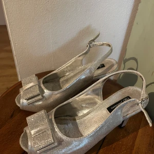 Silvriga klackskor med rosett från Jaclin - Säljer ett par glittriga silvriga klackskor från Jaclin med öppen tå och dekorativ rosett framtill. Skorna har en justerbar rem runt hälen. Köpte dessa på vinted men de passade inte 😣