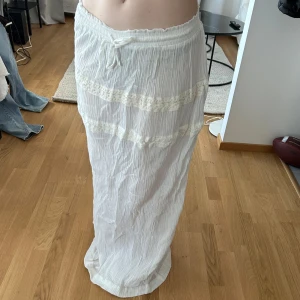 Vit långkjol med spetsdetaljer från PrettyLittleThing - Säljer en vit långkjol från PrettyLittleThing med tunna, vertikala ränder och två breda spetsband som dekoration. Kjolen har resår i midjan och är tillverkad i ett lätt, luftigt material. Perfekt för varma dagar. Linne kjol. Bra att ha på stranden. För stor för mig. Funkar för personer som bär Medium. 