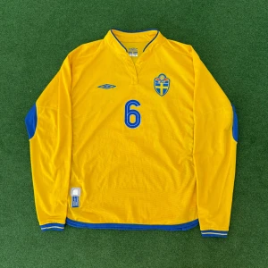 Svenska landslagets långärmade fotbollströja Umbro - Gul långärmad fotbollströja från Umbro med blå detaljer, nummer 6 och svenska landslagets emblem på bröstet. Klassisk V-ringning och blå inslag på sidor och ärmar. Perfekt för dig som älskar svensk fotboll.