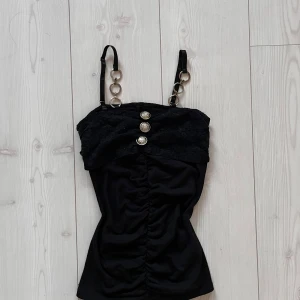 Vintage Topp - Snygg svart axelbandslös topp med dekorativa metallringar på axelbanden och spetsdetaljer upptill. Toppen har en åtsittande passform med rynkade detaljer framtill och tre dekorativa knappar. Toppen är tajt och sitter som en smäck 💓Om ni har frågor så är det bara att kontakta mig. Svarar oftast inom 24h 🥰Står inget märke eller storlek men skulle säga att den sitter som en xs, den är ganska lång men man kan vika upp den utan att det ser konstigt ut. Man kan ha den som en kort klänning också! 💓