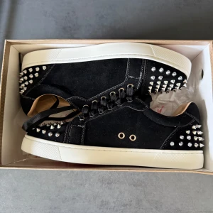 Svarta sneakers med nitar - Snygga svarta sneakers från Christian Louboutin med silverfärgade nitar och röd sula. Skorna har snörning och en rund tå, vilket ger en cool och edgy look. Perfekta för att sticka ut i mängden!