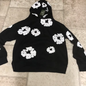 Svart hoodie med vita blommor från Denim Tears - Säljer en svart hoodie från Denim Tears med stora vita blommönster över hela tröjan. Hoodien har en klassisk passform med huva och muddar vid ärmslut och nederkant. Perfekt statement-plagg för dig som vill sticka ut.