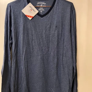 Blå långärmad tröja från Jockey - Säljer en stilren blå långärmad tröja från Jockey med v-ringad hals. Tröjan är tillverkad i mjuk bomull och passar perfekt till vardags. Enkel design med liten broderad logga på bröstet.