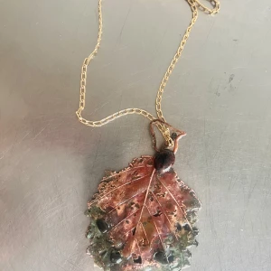 Halsband med lövformat hänge i metall - Unikt halsband med ett stort lövformat hänge i metall med inslag av grönt, koppar och svart. Kedjan är guldig och tunn, vilket ger ett elegant intryck. Hänget har en naturinspirerad design med detaljerade ådror och färgskiftningar.