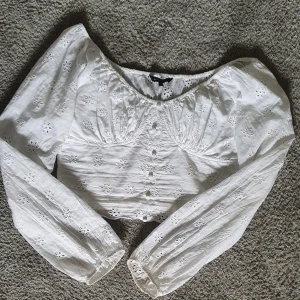 Vit blus med broderade blommor  - Superfin vit blus med broderade blommor och knappar framtill. Blusen har långa puffärmar, croppad passform och smock i ryggen. 