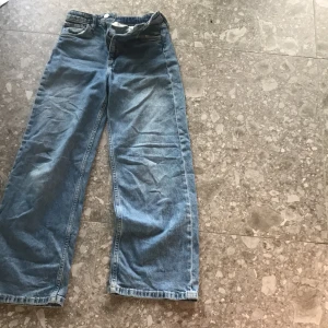 Blå wide jeans från H&M - Säljer ett par blåa wide jeans från H&M med hög midja och justerbar midja. Klassisk femficksmodell med raka, vida ben. Perfekta för en avslappnad och trendig look.