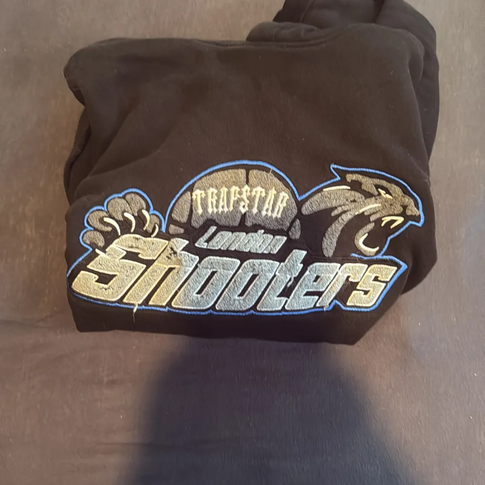 Säljer en svart hoodie från Trapstar med stort broderat London Shooters-motiv på bröstet i blått, vitt och gult. Hoodien har huva, magficka och ribbade muddar. Perfekt för dig som gillar streetwear och sportig stil.. Neuletakit & Villapaidat.