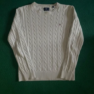 Vit kabelstickad tröja från GANT - Kabelstickad vit tröja från GANT i bomull. Klassisk rund halsringning och diskret broderad logga på bröstet. Perfekt för lager-på-lager och passar till många olika stilar.