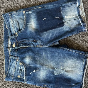 Blå jeansshorts från My Brand - Säljer ett par blå jeansshorts från My Brand med slitna detaljer och färgstänk. Klassisk femficksmodell med knappgylf och patch på bakfickan. Perfekta för en avslappnad stil.