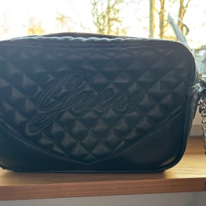 Svart quiltad axelväska från Guess - Säljer en svart quiltad axelväska från Guess med kedjerem och broderad logga på framsidan. Väskan har ett ljust innerfoder med mönster och flera fack för enkel organisering. Perfekt för att bära dina viktigaste saker.