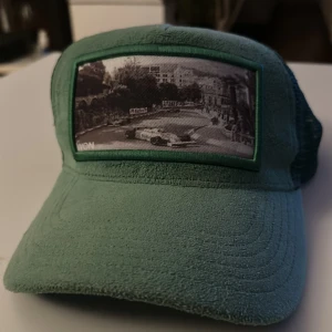 Grön keps med svartvit bild - Säljer en grön keps med nät på baksidan och en svartvit bild framtill som motiv. Kepsen har böjd skärm och gröna detaljer runt bilden. Perfekt för dig som gillar unika accessoarer.