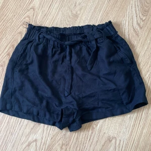 Svarta shorts med knyt från KappAhl - Svarta shorts från KappAhl med resår i midjan och knytband framtill. Modellen har två sidofickor och är tillverkad i ett mjukt tyg som känns skönt mot huden. Perfekta för varma dagar.
