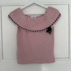 Ljusrosa stickad topp med volangkrage - Supersöt ljusrosa stickad topp med bred volangkrage och svart dekorativ kant. En svart rosett sitter på ena sidan av kragen för extra detalj. Perfekt för dig som vill ha något unikt och gulligt i garderoben. Prislapp kvar