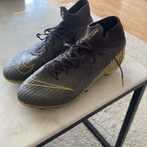 Nike Mercurial Superfly 6 elite AG - Säljer ett par Nike Mercurial Superfly fotbollsskor i svart med gula detaljer och mönster. Skorna har högt skaft och snörning, samt dobbar för spel på gräs. Perfekta för dig som vill ha stilrena och snabba skor på planen.