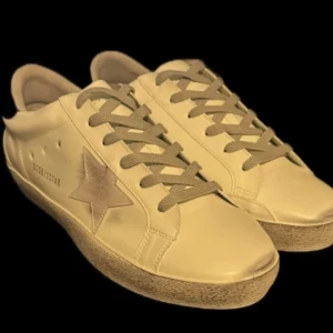 Golden Goose Superstar sneakers i beige - Golden Goose Superstar sneakers i beige med ikonisk stjärndetalj på sidan och svart hälflik med logga. Skorna har snörning och en slitstark gummisula. Perfekta för en avslappnad och trendig look. Priset är inte fast skriv vid frågor (skorna är rätt så slitna med dem funkar)