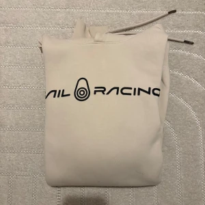 Sail Racing - Säljer en snygg beige hoodie från Sail Racing med svart logga på bröstet. Tröjan har en klassisk känguruficka och justerbar huva med dragsko. Perfekt för en avslappnad stil. Storlek S. Kvitto finns